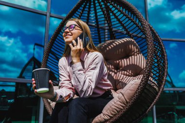 Tarz sahibi genç bir kadın rahat bir sandalyede rahatlar, kahvenin tadını çıkarır ve telefonda sohbet eder, açık mavi gökyüzünün altında, doğa tarafından çevrelenmiş, keyifli bir anın tadını çıkarır.
