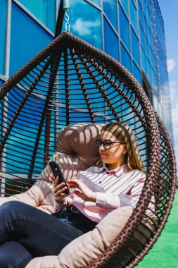 Genç bir kadın şık bir sallanan sandalyede rahat bir şekilde oturuyor, akıllı telefonuna dalmış, çağdaş bir şehir zemininde, boş zamanlarını ve modern yaşamını sergiliyor..