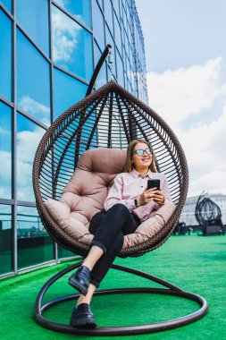 Modaya uygun bir kadın, ferah bir terasta rahat bir sallanan sandalyede, modern mimarinin ve açık mavi gökyüzünün ortasında akıllı telefonuyla mesajlaşıyor.