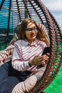 Genç bir kadın modern bir sallanan sandalyede rahatlar, akıllı telefonunu açık havada hareketli gölgeler oluşturan parlak güneş ışığı altında mutlu bir şekilde kullanır.