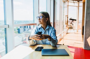 Genç bir profesyonel, şık ve modern ofisinde akıllı bir telefon kullanırken başarılarını yansıtıyor ve enerjik bir çalışma ortamında verimliliğini gösteriyor.