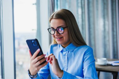 Moda gözlüklü şık bir genç kadın, akıllı telefonuyla günümüzün en son iletişim ve teknoloji eğilimlerini yansıtıyor.