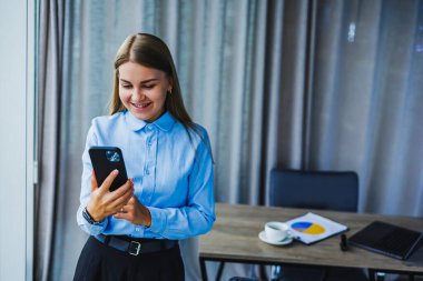 Modern bir ofiste genç bir kadın akıllı telefonunu kullanırken gülümsüyor ve günümüz toplumunda sağlıklı bir iş hayatı dengesini korumanın önemini vurguluyor.
