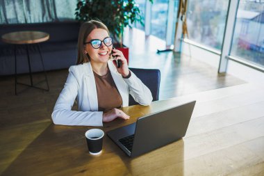 Modern bir ofiste, kendine güvenen bir iş kadını dizüstü bilgisayarı üzerinde çalışırken üretkenliğini, zarafetini ve profesyonelliğini sergileyerek dinamik bir telefon görüşmesi yapar.