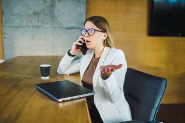 Şık beyaz ceketli odaklanmış bir iş kadını modern bir masada telefonla konuşuyor. Dinamik çalışma alanında profesyonelliğini ve kararlılığını gösteriyor.