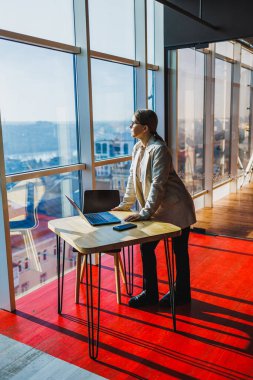 Kendine güvenen bir iş adamı modern bir ofiste şık bir masada duruyor geniş pencerelerden geniş şehir manzarasına bakıyor, kurumsal odak ve hırsı somutlaştırıyor.