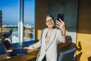 Neşeli ve kendinden emin genç bir profesyonel şık, çağdaş bir ofis ortamında arka planda çarpıcı bir şehir manzarasıyla gururla gülümsemesini gösteren bir selfie çekiyor.