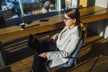 Kendini işine adamış ve hırslı bir kadın şık ofis koltuğunda oturmuş laptopuyla çalışırken üretkenliğini ve yaratıcılığını arttıran nefes kesici şehir manzarasının tadını çıkarıyor.