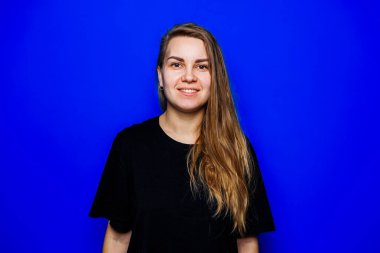 Dalgalı saçları olan neşeli genç bir kadın mavi bir arka planda kendine güven yayıyor, bulaşıcı gülümsemesiyle büyüleyici bir pozitiflik ve çaba gerektirmeyen bir tarz yayıyor.