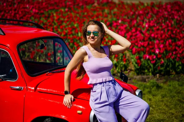 Güneş gözlüklü şık bir kadın canlı kırmızı bir klasik arabanın yanında poz veriyor, göz kamaştırıcı pembe lale çiçeklerinin ortasında, resim gibi bir sahne yaratıyor.