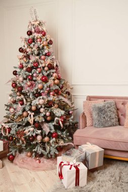 Süs ve kurdelelerle süslenmiş güzel dekore edilmiş bir Noel ağacı rahat bir kanepenin yanında duruyor ve altında hediyeler olan bayram havası yaratıyor..