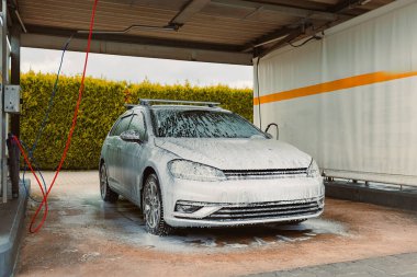 Kalın sabun köpüğüyle kaplanmış, iyice yıkanmış bir araba, parlak bir dış görünüşü gösteriyor. Korunaklı yıkanma alanı canlı yeşil bir zemin ile zenginleştiriliyor ve canlı bir sahne yaratıyor.