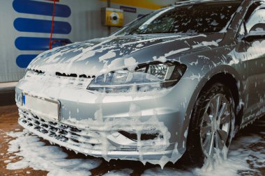 Araba yıkamada köpüklü sabunla kaplanmış gümüş renkli bir araba, araç görünümünü korumak için temizleme sürecini gösteriyor.