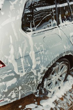 Yıkama işlemi sırasında kalın sabun köpüğüyle kaplı parlak gümüş bir arabanın yakın çekim görüntüsü, parlak kıvrımlarını ve çok sayıda küçük kabarcıkları sergiliyor.