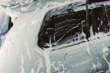 Bu görüntü, araba bakımının titiz bir şekilde temizlenmesi için cam ve gövdeyi kaplayan yoğun sabun köpüğüyle yıkanan bir arabanın yakın çekimini gösteriyor.