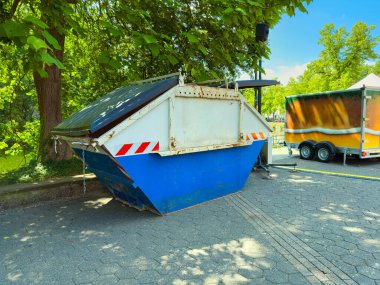 Mavi ve beyaz atık konteynırı açık havada, ağaçlarla kısmen gölgelenmiş, onu saran yemyeşil çevreyle canlı bir kontrast oluşturuyor.