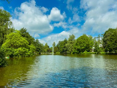Huzurlu bir parkta, sakin bir göl yemyeşil ve açık mavi gökyüzüyle çevrili çarpıcı bir çeşmeyi yansıtır. Doğa severlerine rahatlama için ideal bir kaçış sunar.