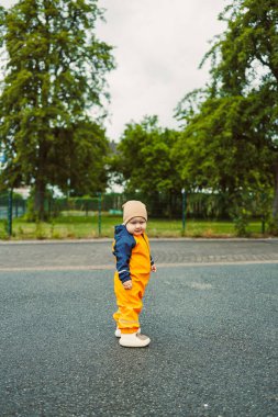 Canlı turuncu yağmurluk giymiş şirin bir çocuk, arka planda yeşillik olan bir yolda neşeyle duruyor, çocukluk macerasının özünü yakalıyor.