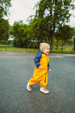 Canlı turuncu yağmurluk ve rahat bir bere giyen mutlu bir çocuk yeşil ağaçlarla çevrili yumuşak asfalt bir yolda neşeyle koşar, çocukluk masumiyetini somutlaştırır..
