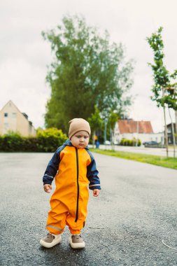 Parlak turuncu yağmurluk giymiş neşeli ve neşeli bir çocuk, sakin bir açık hava ortamını zevkle keşfeder, çocukluk macerasının, merakının ve neşesinin saf özünü yakalar