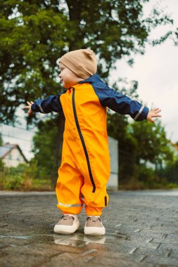 Turuncu yağmurluklu ve rahat şapkalı neşeli bir çocuk yağmurda neşeyle oynuyor, neşe ve özgürlük saçıyor, eğlenceli ve maceralı bir günün büyülü anlarını kucaklıyor.