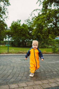 Yağmurlu bir günde parlak turuncu bir yağmurluk giymiş neşeli bir çocuk neşeli bir şekilde yeşil alanda yürüyor ve onları çevreleyen doğanın güzelliğine dalıyor.