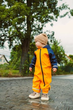 Parlak sarı bir yağmurluk ve kalın bir şapka giymiş hoş bir çocuk, etrafı yemyeşil yeşilliklerle çevrili yağmurlu bir sokağı neşeyle keşfeder.