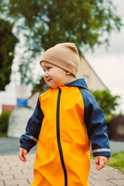 Parlak sarı yağmurluk ve rahat bir şapkayla neşeli bir çocuk bulutlu bir günde açık havada gezer, tasasız çocukluk macerası ve eğlencenin özünü somutlaştırır