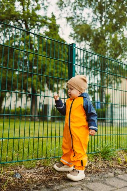 Parlak turuncu bir yağmurluk ve rahat bir şapka giymiş, güneşli bir günde yemyeşil bir çitin yanında gezen sevimli bir çocuk.