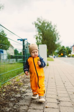 Bulutlu bir günde, parlak turuncu bir yağmurlukla neşeli bir çocuk ahşap bir çitin kenarında geziniyor, merak ve heyecanla doğayı keşfediyor.