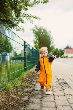 Parlak turuncu yağmurluk giymiş neşeli genç bir çocuk, bereketli yeşilliğin ve manzaralı bir yolun ortasında sonsuz bir merak göstererek, açık havada mutlu bir şekilde gezer.