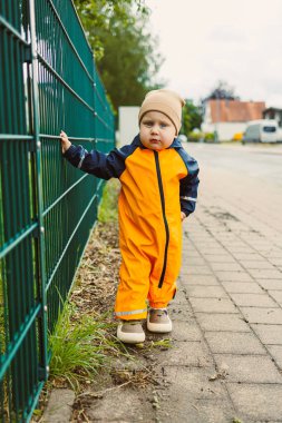 Turuncu yağmurluklu ve şapkalı neşeli bir çocuk yeşil bir çitin kenarındaki kaldırımı keşfeder, açık havada neşeli anlar gösterir, hafif yağmurda bile, güzel anılar yaratır.