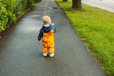 Parlak turuncu yağmurluklu çekici bir çocuk, çocukluk, doğa ve keşif temaları için ideal yemyeşil bir yolda mutlu bir şekilde yürüyor.