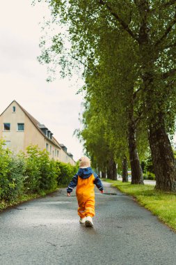 Canlı yağmurluk giymiş neşeli bir çocuk, bereketli bir yağmurun keyfini çıkardıktan sonra yemyeşil ve güzel evlerle çevrili ıslak bir patika boyunca mutlu bir şekilde yürür.