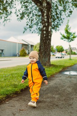 Canlı turuncu bir yağmurluk giymiş neşeli küçük bir çocuk, bulutlu bir günde, ağaçlı bir kaldırımda mutlu bir şekilde geziniyor, muhteşem açık havayı kucaklıyor.