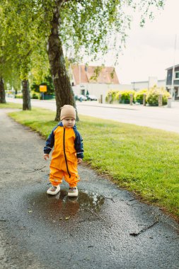 Parlak turuncu yağmurluk giymiş neşeli, hevesli bir çocuk neşe içinde bir su birikintisinde duruyor, yemyeşil yemliklerle çevrili sıcak, güneşli bir günde su sıçratmaya hazırlanıyor.