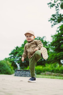 Neşeli genç bir çocuk, şık bir şekilde giyinmiş, neşe dolu yeşil bir parkı keşfediyor, doğanın güzelliğine dalmış, merak ve keyif saçıyor.