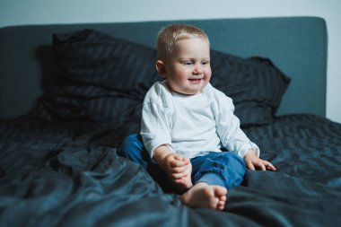 Neşeli bir bebek mutluluk saçar parlak bir gülümsemeyle, yumuşak yastıklarla çevrili rahat bir yatakta oturur, sıcak ve davetkar bir masumiyet ortamı yaratır.