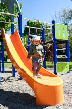 Meraklı bir çocuk, bu güneşli günde eğlenmek ve eğlenmek için parlak bir turuncu kaydırağın üzerinde durur ve diğerlerini de katılmaya davet eder.