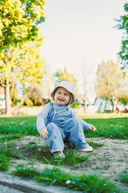 Aydınlık, güneşli bir günde, mutlu genç bir çocuk dışarıda bir parkta neşeli bir şekilde oynuyor, çimenlerin üzerinde oturuyor, yemyeşil ağaçlarla çevrili, ve masum, neşeli bir yüz ifadesi sergiliyor.