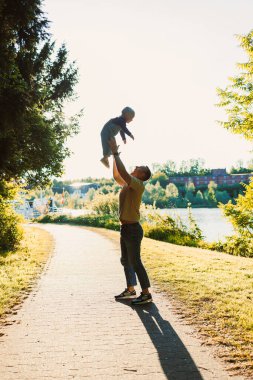 Nehir kıyısında güneşli güzel bir günde, sevgi dolu bir baba, doğanın çarpıcı güzelliğinin ortasındaki keyifli bağlarını göstermek için, küçük oğlunu sevinçle havaya kaldırır.