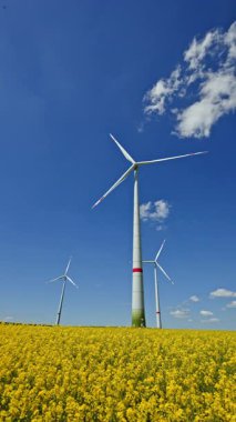 Kırsaldaki mavi gökyüzüne karşı sarı bir tarlada rüzgar türbini. Avrupa 'daki tarım arazilerinde fırtına bulutları olan rüzgâr tarlası. Rüzgar santrali elektrik üretiyor.