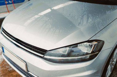 Parlak, gümüş renkli bir araba kaputu, iyice yıkandıktan sonra su damlacıklarıyla parlıyor. Parlak yüzeyi ve iyi tasarlanmış farları sergiliyor. Otomotiv görüntüleri için mükemmel..