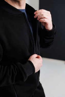 Gösterişli, siyah fermuarlı bir ceket giymiş şık bir adam minimalist bir arka plan önünde kıyafetlerini düzeltirken görülüyor. Modern gündelik moda eğilimlerini mükemmel bir şekilde gözler önüne seriyor.