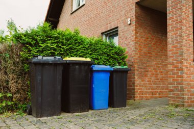 Kırmızı tuğlalı bir evin önündeki renkli atık kutuları, sürdürülebilir, çevre dostu topluluklar oluşturmak için verimli atık yönetimi ve geri dönüşümün önemini vurguluyor