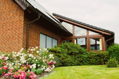 Şık tuğlalı dış görünüşü ve geniş pencereleri olan göz kamaştırıcı modern bir ev, canlı çiçekler ve yemyeşil ile çevrili, büyüleyici bir açık hava ortamı yaratıyor.