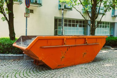 Yeşil ağaçlar ve modern binalar tarafından kucaklanan bir kaldırım taşı caddesinde duran turuncu bir çöp konteyneri, kentsel gelişimi ve dönüşümü sembolize ediyor.