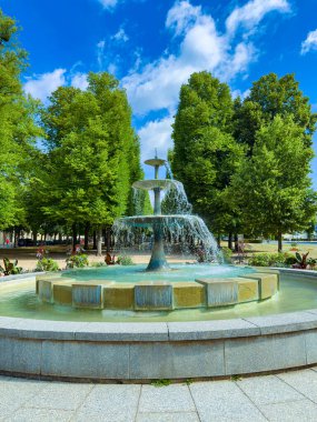 Bereketli ağaçlar ve mavi gökyüzü ile çevrili sakin bir parkta güzel bir çeşme, doğanın rahatlaması ve takdir edilmesini teşvik eden sakin bir atmosfer yaratır.