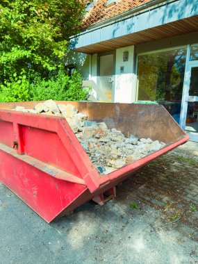 Girişin yanında, açık, güneşli bir gökyüzünün altında yemyeşil bir güzellikle vurgulanan çeşitli yapı atıklarıyla dolu taşmış kırmızı bir çöp konteyneri var.