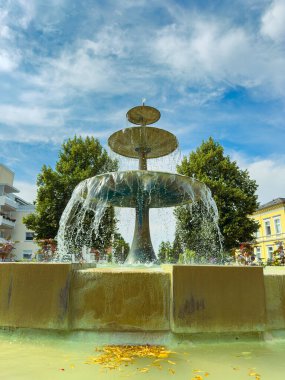 Çoklu katmanlı güzel bir çeşme, canlı bir yeşillik zeminine ve canlı bir mavi gökyüzüne karşı nazikçe su dökerek, huzurlu bir şehir atmosferi yaratıyor..